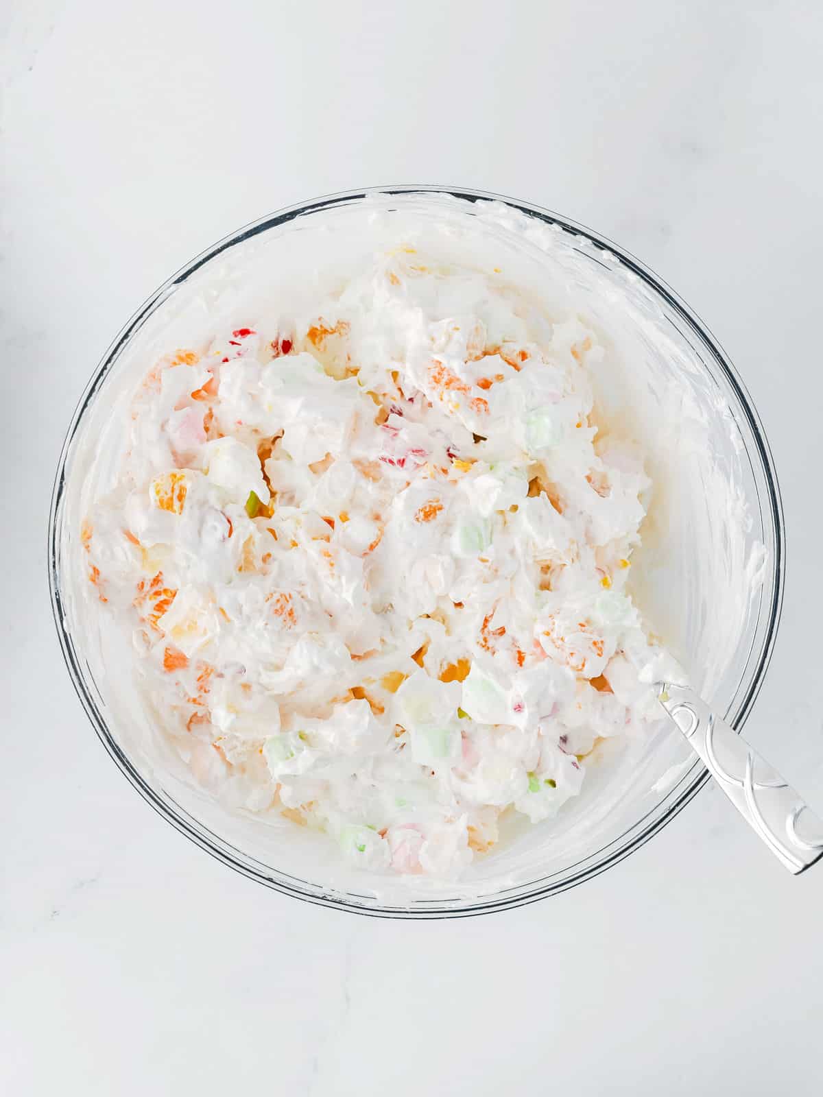 A glass bowl filled with a creamy fruit salad mixture, containing whipped topping, fruit pieces, and mini marshmallows, with a metal spoon resting inside.