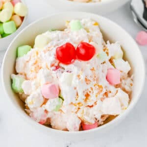 A white bowl filled with ambrosia salad, containing whipped cream, colorful mini marshmallows, fruit pieces, and topped with two maraschino cherries.