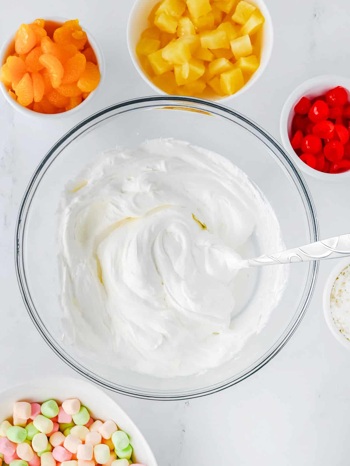 A glass bowl of whipped topping with a spoon, surrounded by bowls of mandarin oranges, pineapple chunks, cherries, shredded coconut, and colorful mini marshmallows.
