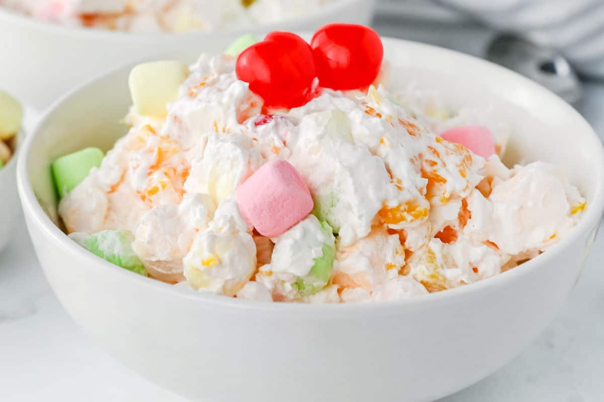 A bowl of ambrosia salad topped with two maraschino cherries, featuring whipped cream, fruit, and colorful mini marshmallows.