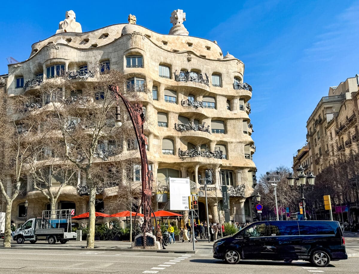 A unique stone building designed by Gaudi with wavy balconies and roof sculptures stands on a busy city street, where cruise shore excursions often stop to let visitors admire the architecture amid cars, trees, and people on a sunny day.