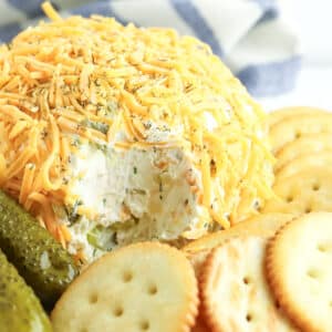 A cheese ball covered with shredded cheddar, surrounded by round crackers and pickle spears on a white surface.