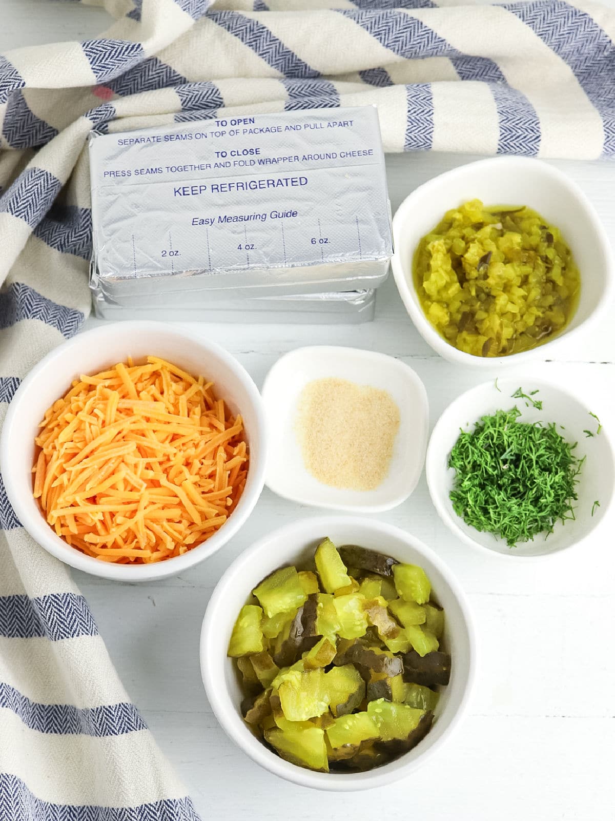 Five small bowls contain shredded cheddar cheese, chopped pickles, chopped fresh herbs, relish, and a seasoning powder. A package of cream cheese is in the background on a striped cloth.