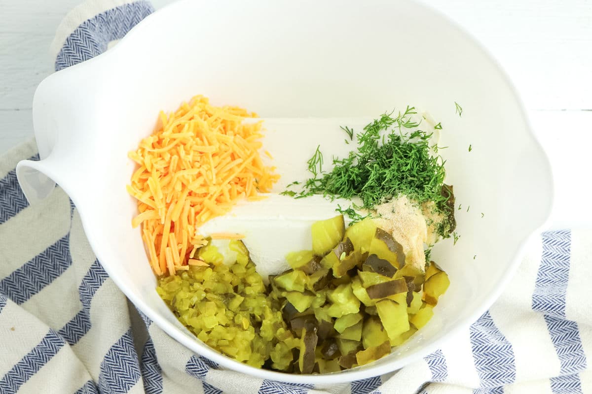 A white mixing bowl contains shredded cheddar cheese, cream cheese, chopped pickles, dill, and seasonings, placed on a striped cloth.