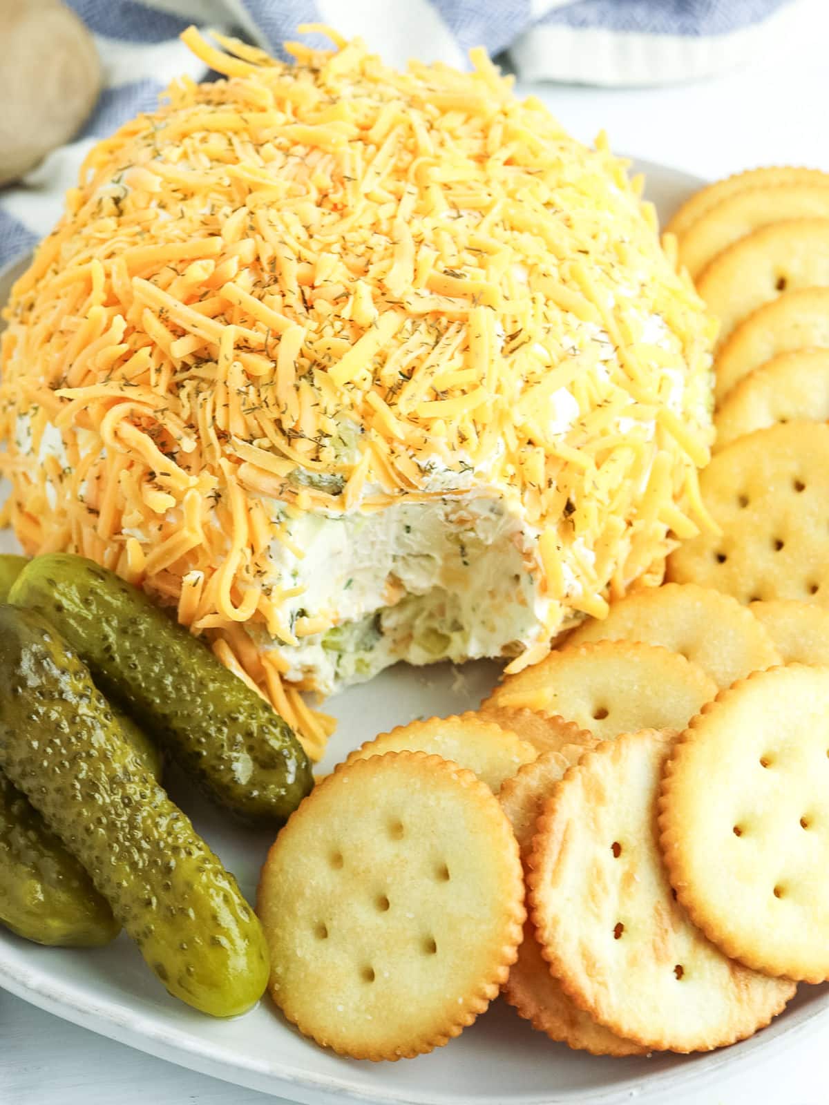 A cheese ball coated with shredded cheese, served on a plate with round crackers and pickles.