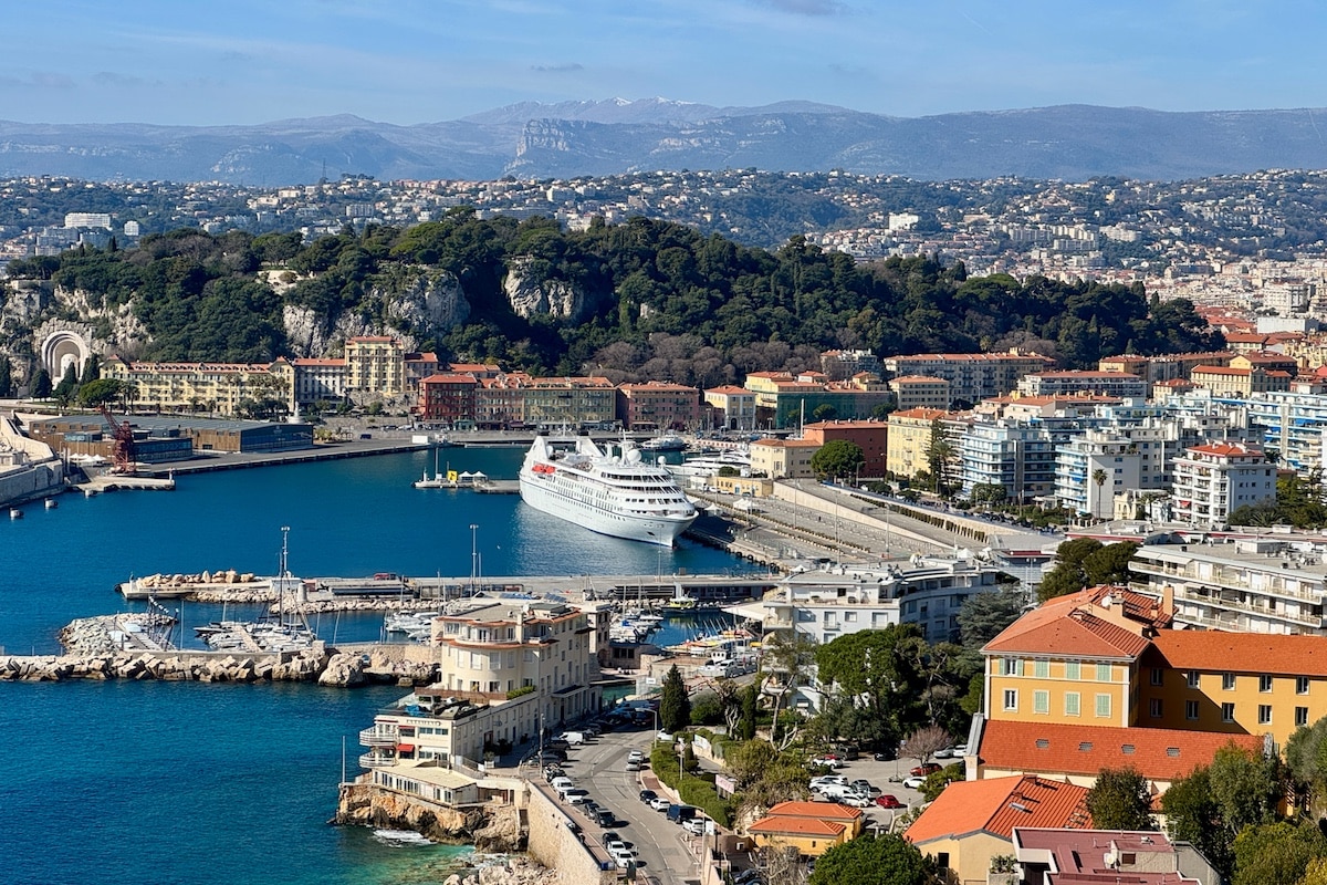 The Windstar Star Legend cruise ship docked at a harbor in a Nice, France, with colorful buildings, a marina, and hills beyond&mdash;an inviting scene for cruise shore excursions under a clear sky.