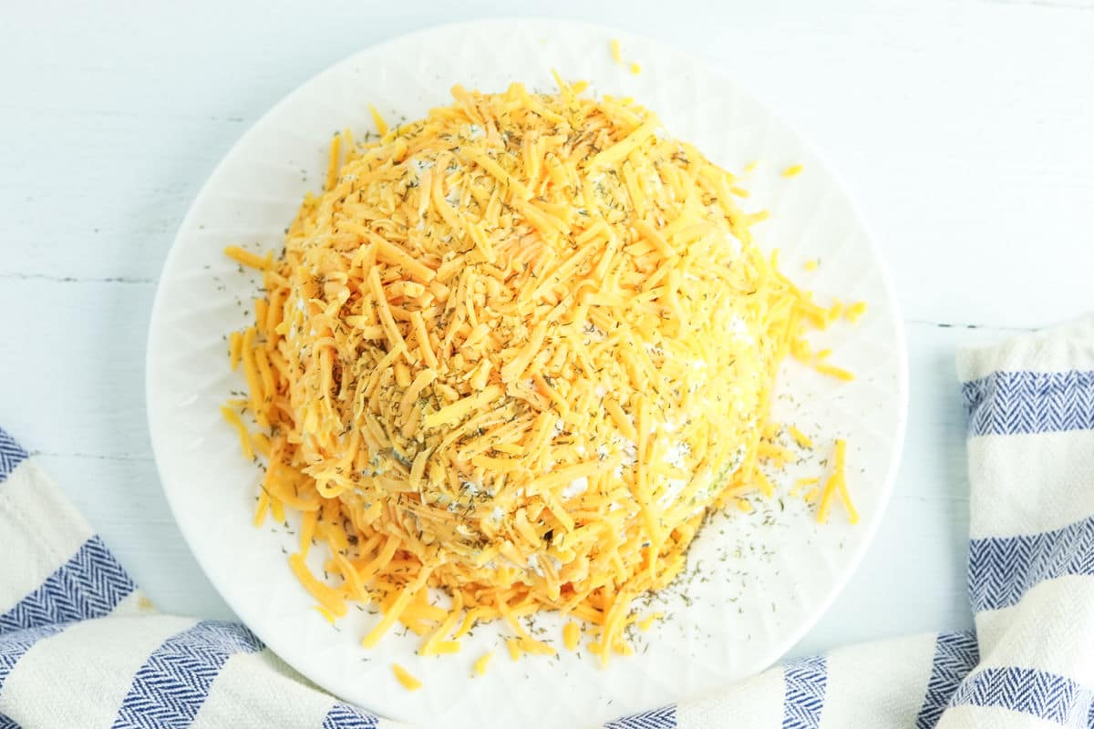 A ball-shaped cheese spread covered in shredded cheddar cheese and herbs on a white plate, with a striped cloth nearby.
