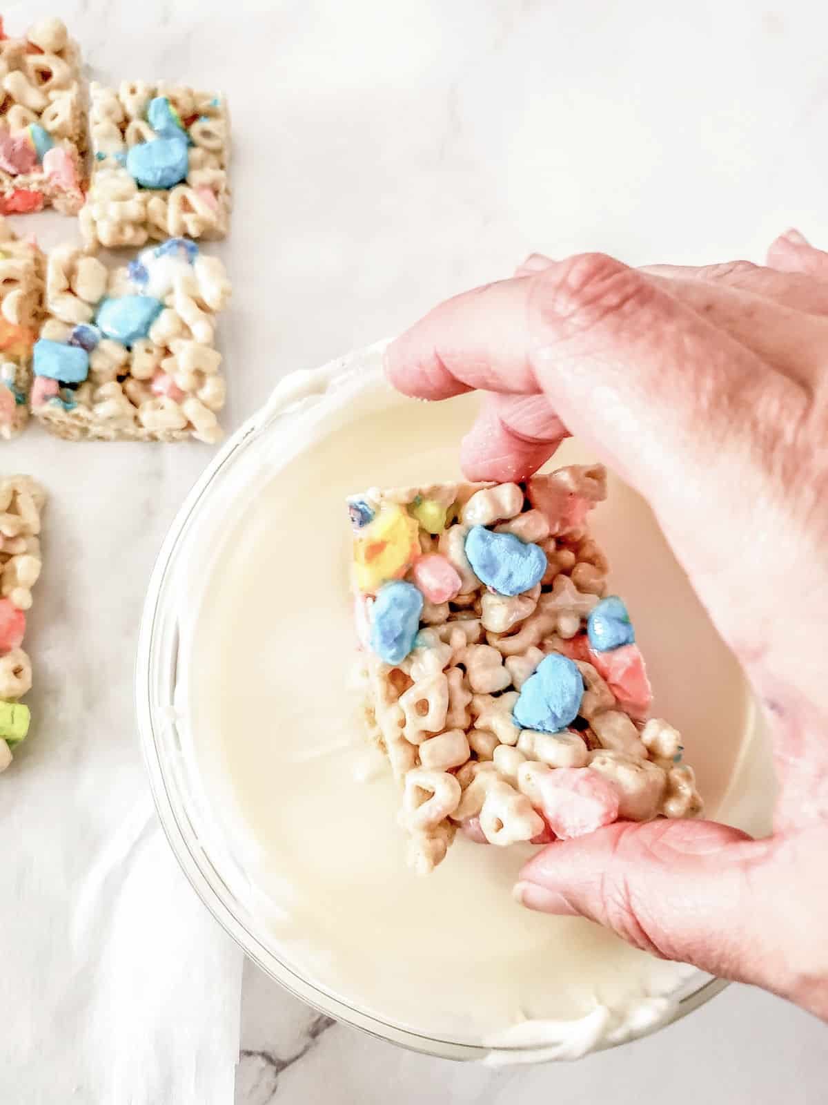 A hand dips a square marshmallow cereal treat into a bowl of white melted chocolate, with more treats on parchment in the background.