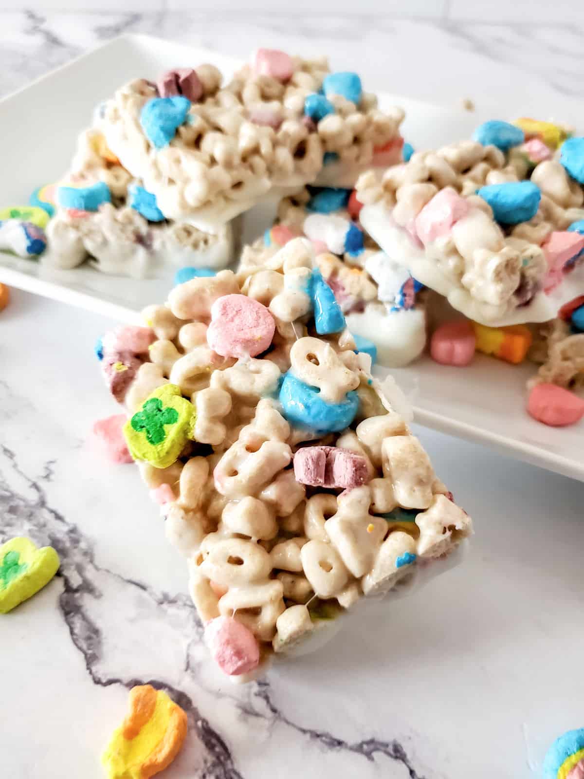 A close-up of cereal bars made with colorful marshmallow cereal pieces, stacked on a white plate on a marble surface.