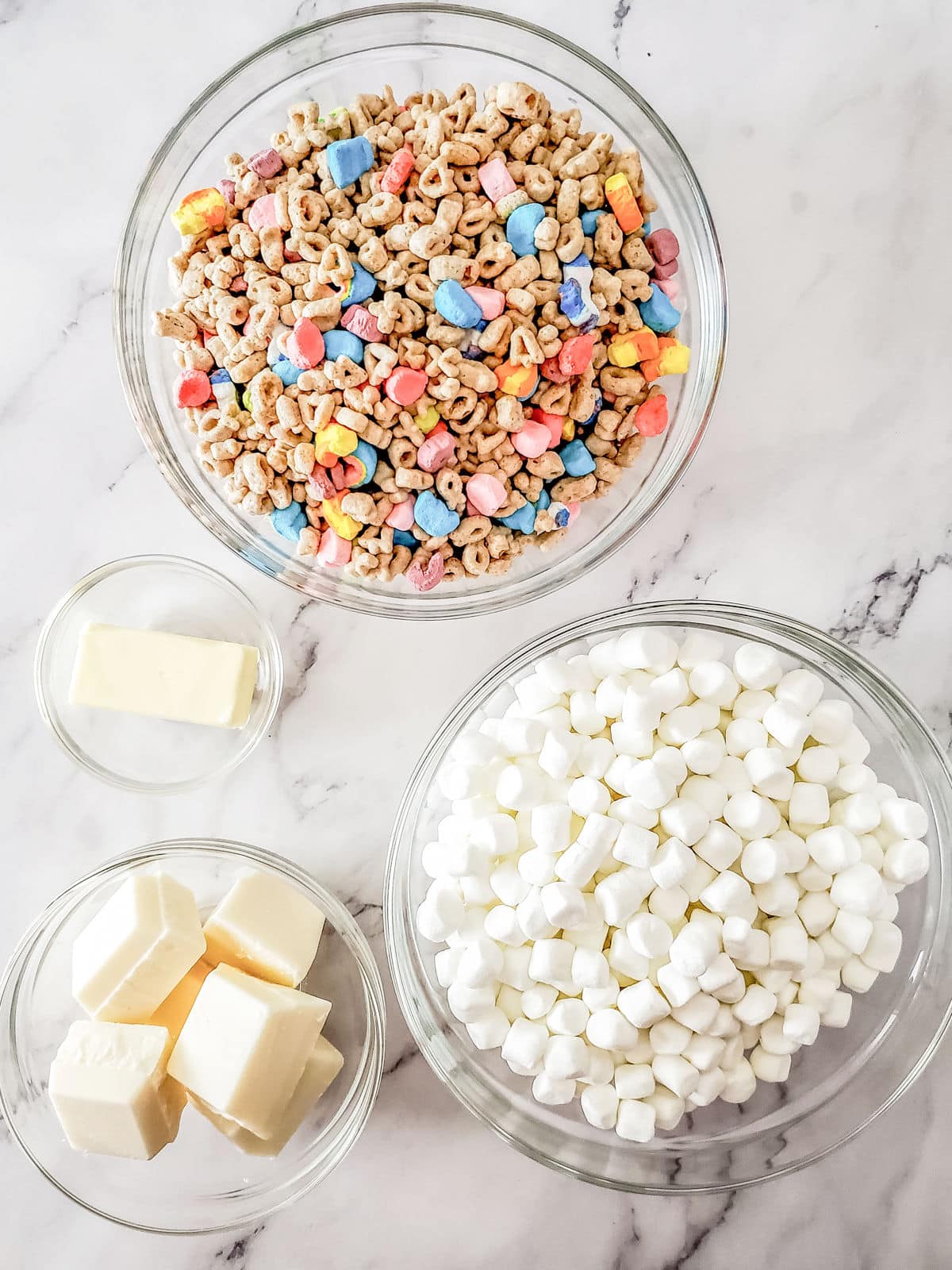Four glass bowls containing colorful cereal, mini marshmallows, butter, and white candy melts are arranged on a white marble surface.