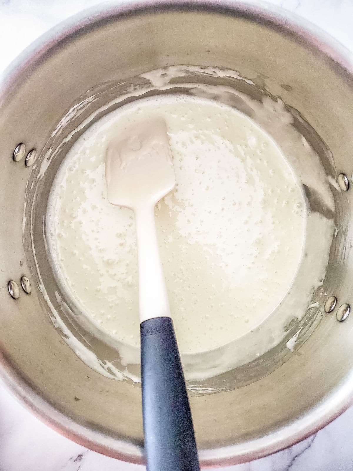 A saucepan containing a creamy white mixture being stirred with a white spatula, set on a white marble surface.