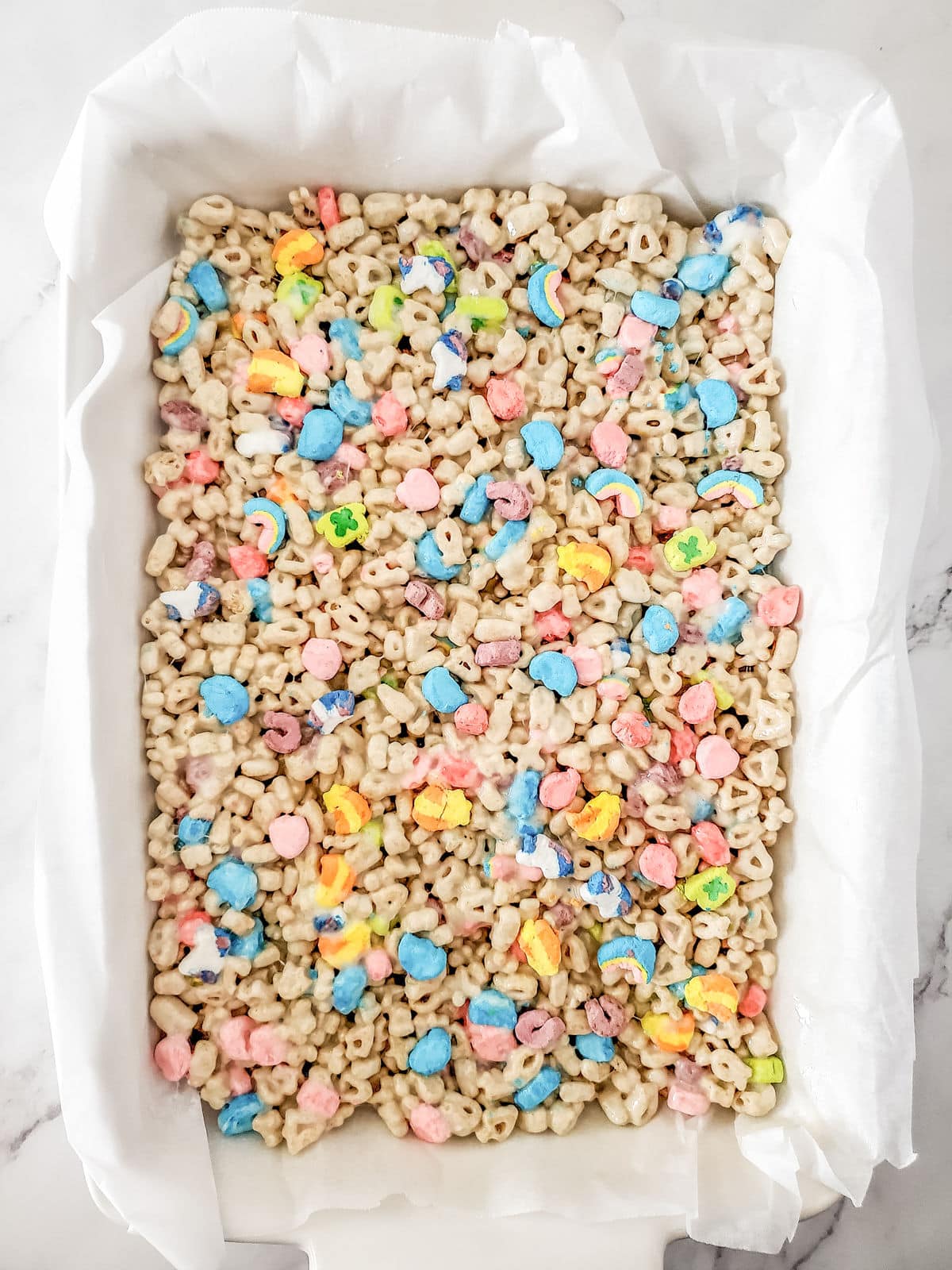 A rectangular baking dish lined with parchment paper holds a mixture of cereal pieces and colorful marshmallow shapes, spread evenly.