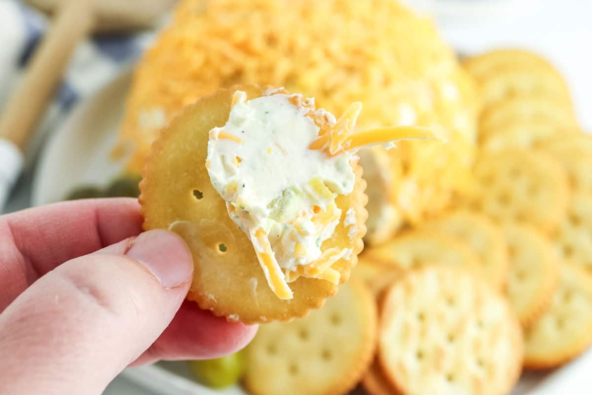 A hand holds a round cracker topped with cheese ball spread; more crackers and a cheese ball are visible on a plate in the background.