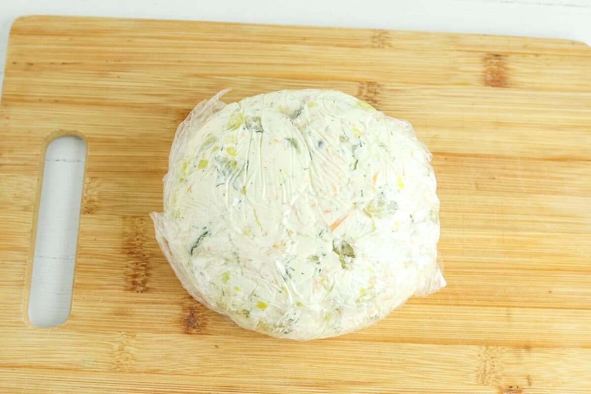 A ball of cheese wrapped in plastic wrap sits on a wooden cutting board.