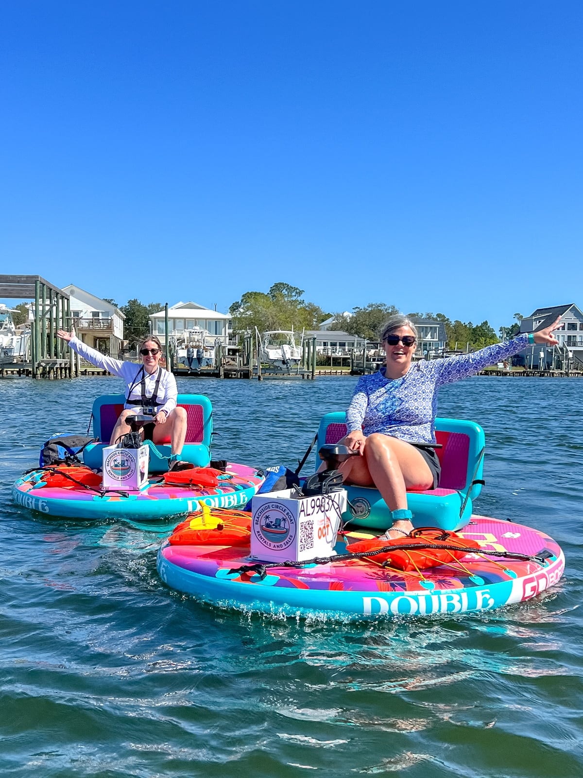 Two women sit on colorful circular paddle boats on calm water, arms outstretched, with houses and docks visible in the background under a clear blue sky.