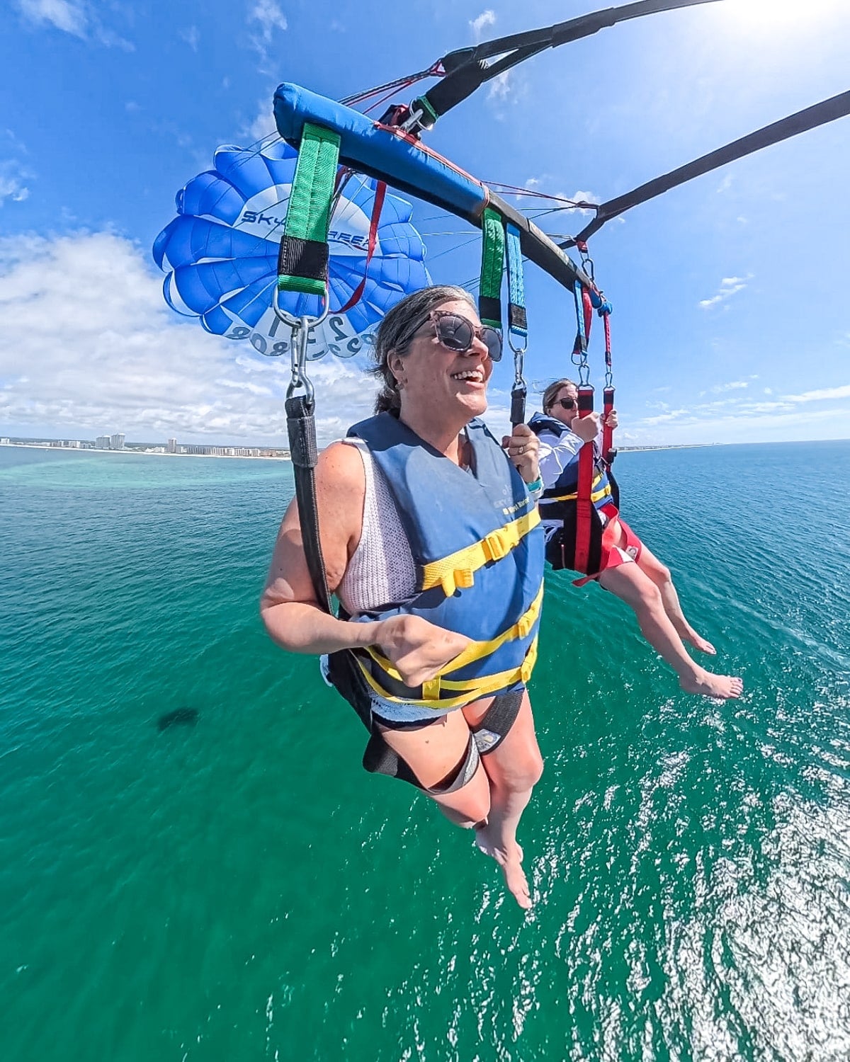 Two people wearing life jackets parasailing over clear blue ocean water, with a shoreline and buildings visible in the distance under a partly cloudy sky.