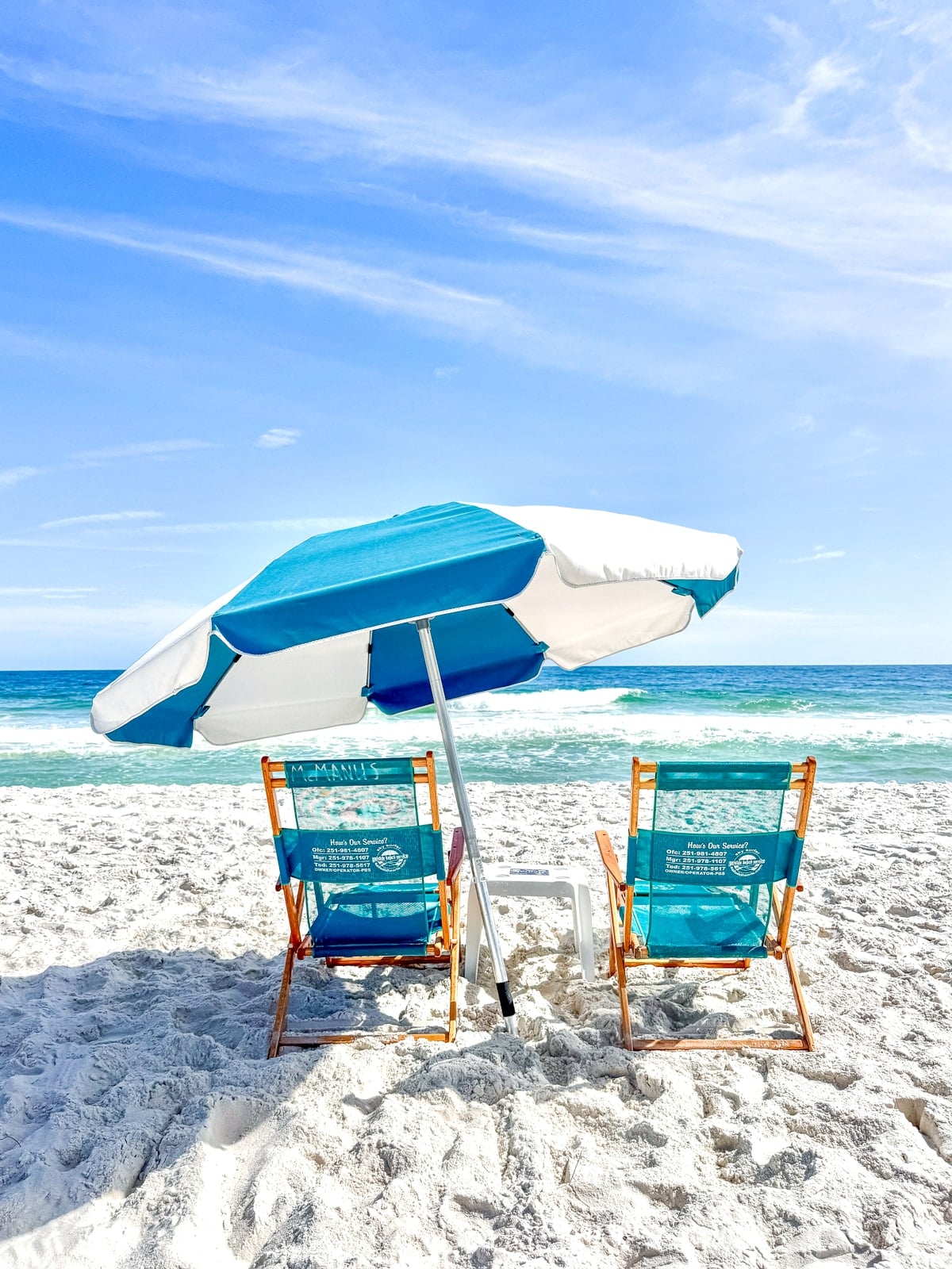 Two empty beach chairs and a blue-and-white umbrella are set up on white sand facing the ocean under a clear blue sky.