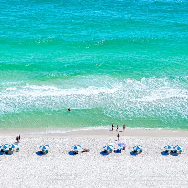 Aerial view of people on a sandy beach with blue and white umbrellas near turquoise ocean waves.