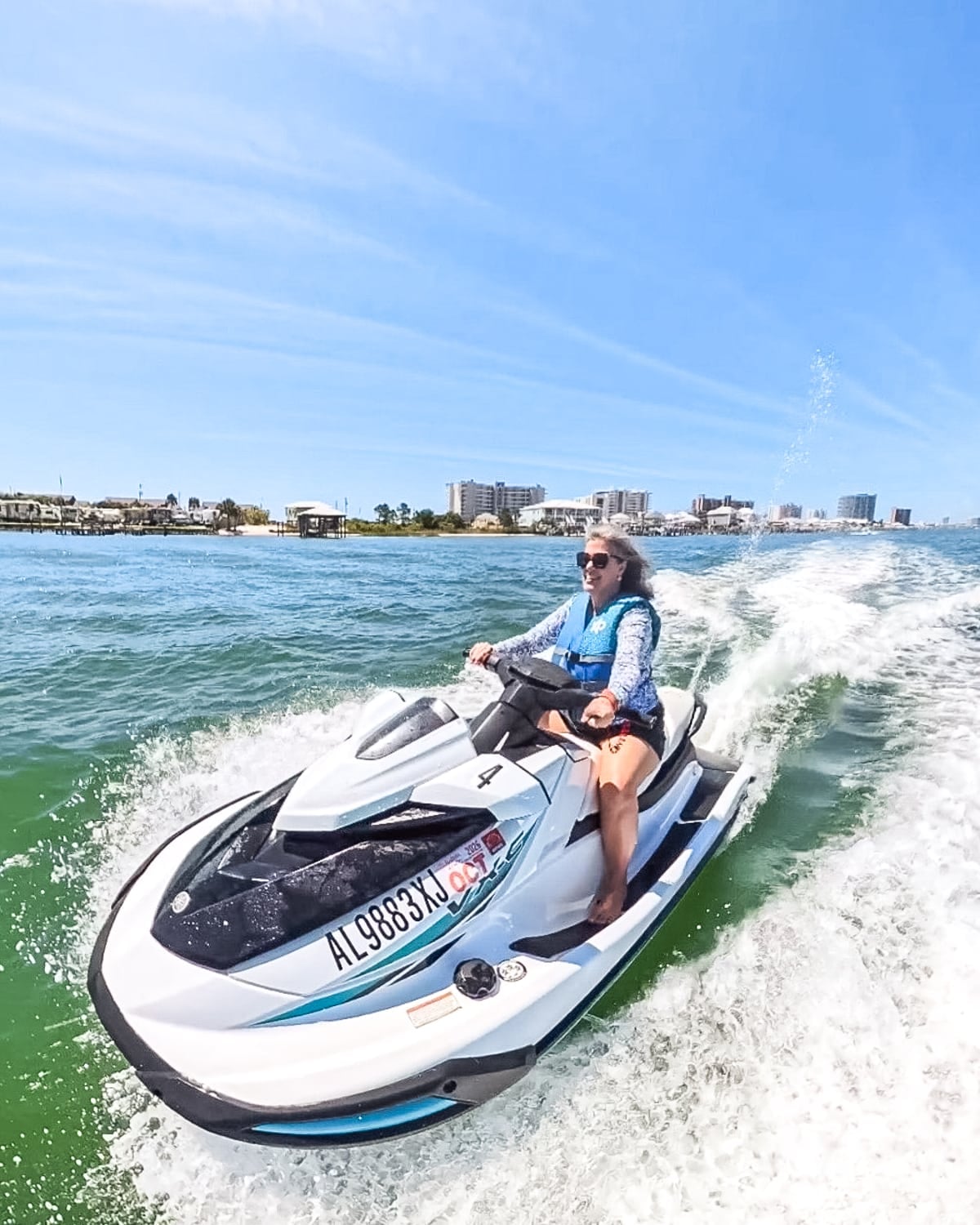 A person wearing sunglasses and a life jacket rides a white jet ski on the water near a coastal city under a clear blue sky.