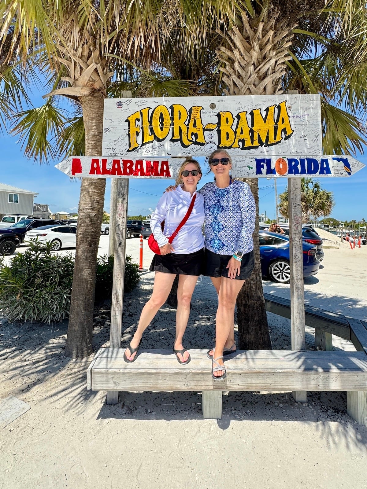 Two women stand under a &ldquo;Flora-Bama&rdquo; sign between Alabama and Florida state markers on a sunny beach, with palm trees, sand, and parked cars in the background.