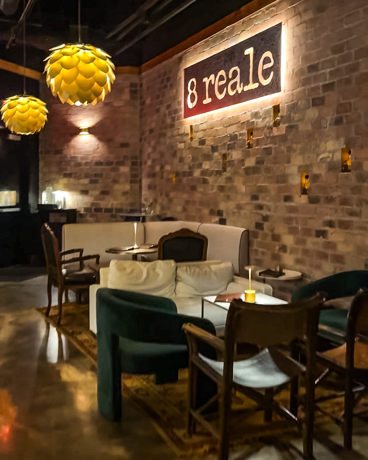 A cozy lounge with brick walls, warm lighting, assorted chairs and sofas, and a sign reading "8 reale" mounted on the wall.