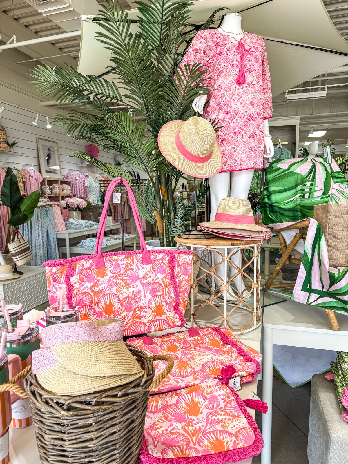 Display of pink and green resort wear and accessories, including hats, bags, and clothing, arranged on tables and mannequins in a brightly lit retail store.