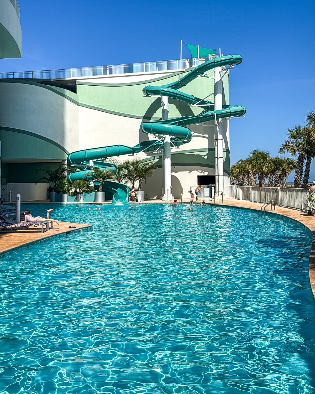 Large outdoor pool with clear blue water and a tall, twisting green water slide attached to a modern building; a few people are swimming and relaxing by the poolside.