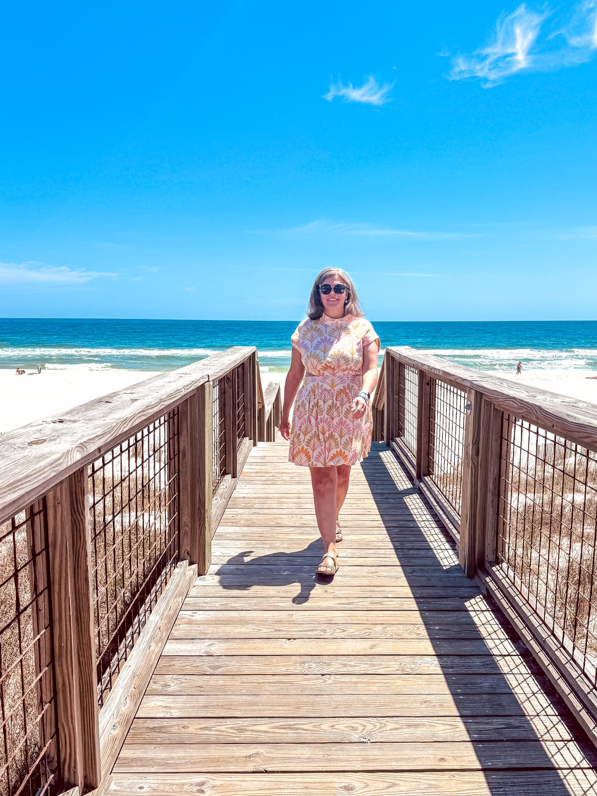 A woman in a patterned dress walks on a wooden boardwalk toward a sandy beach and blue ocean under a clear sky.