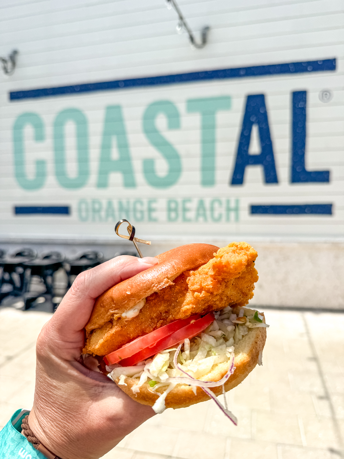 A hand holds a fried chicken sandwich with lettuce, tomato, and onion in front of a wall sign reading "COASTAL ORANGE BEACH.