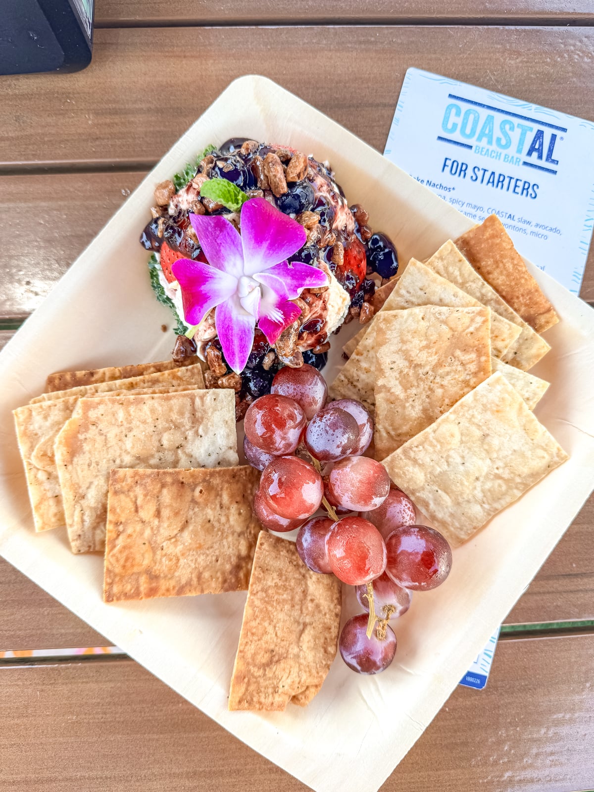 A square plate with crackers, red grapes, and a cheese ball topped with dried fruit, nuts, and a purple flower, placed on a wooden table next to a Coastal Beach Bar menu.