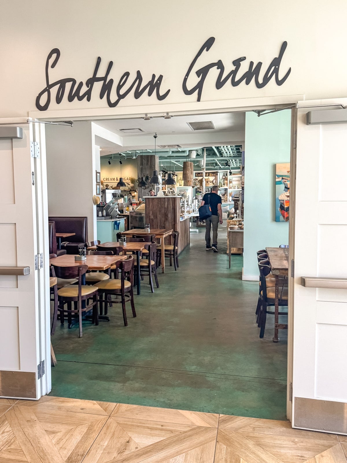 Entrance to a cafe named "Southern Grind" with tables and chairs inside, a person walking toward the counter, and shelves displaying various items.