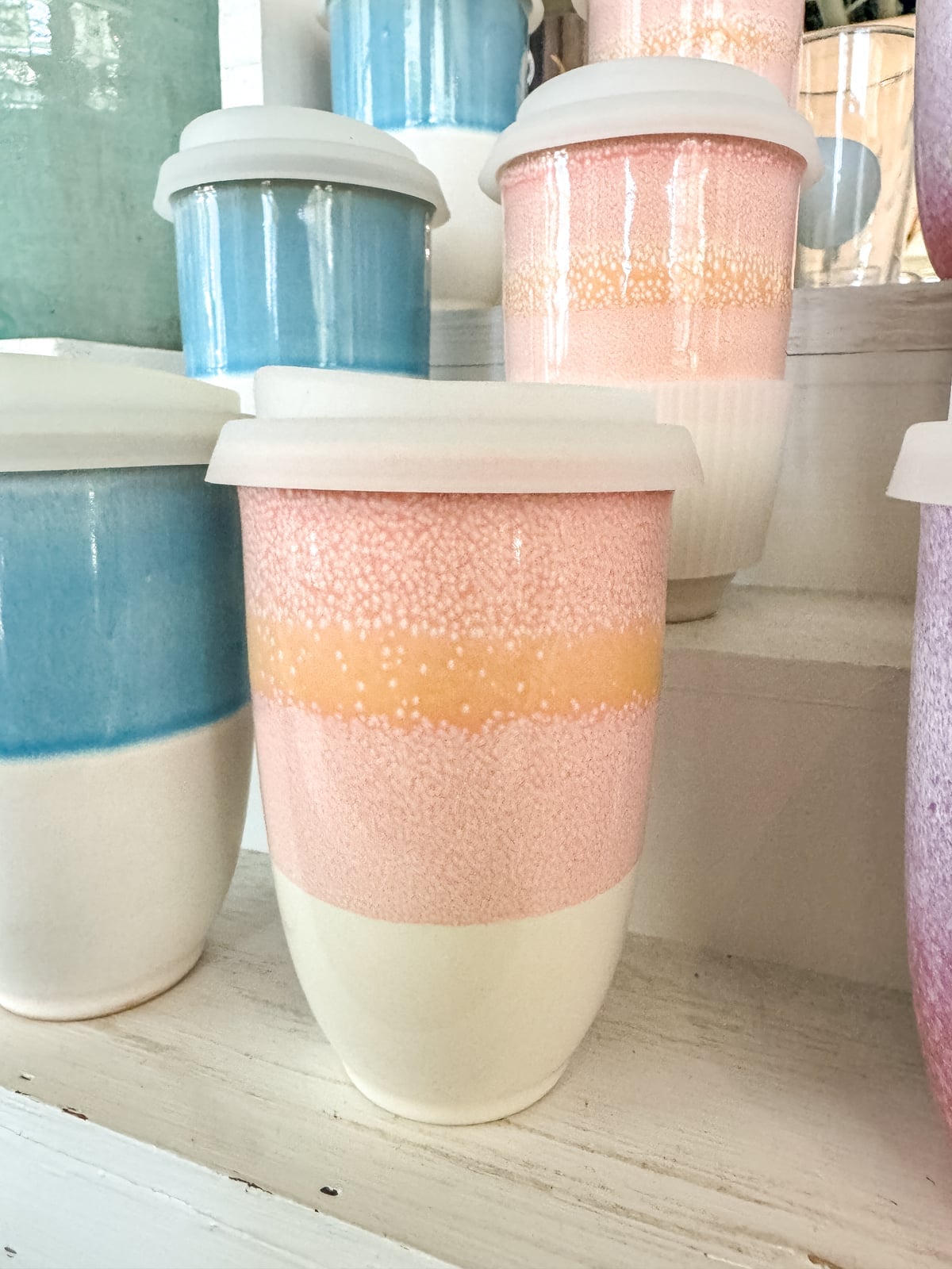 Ceramic cups with white lids are displayed on shelves. The cups feature pastel colors, including pink, blue, and peach, with a gradient or speckled design.