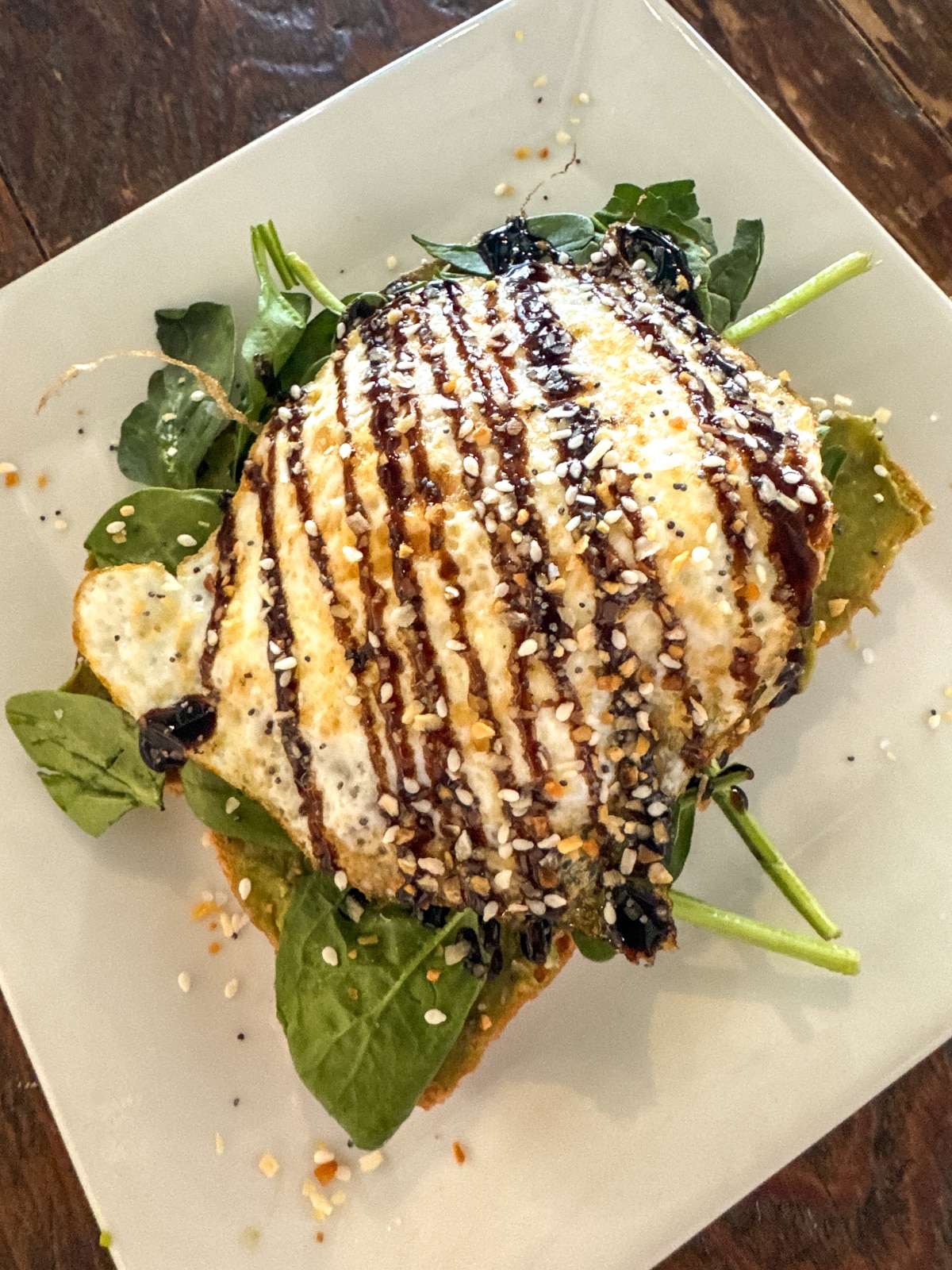 Open-faced sandwich topped with greens, a fried egg, sesame seeds, and a drizzle of balsamic glaze on a square white plate.