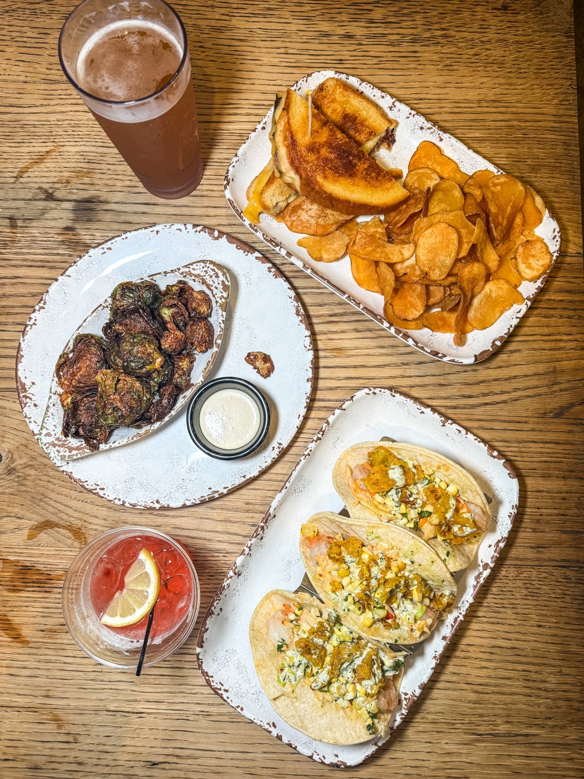 A wooden table set with a grilled cheese sandwich and chips, three fish tacos, fried Brussels sprouts with dip, a pint of beer, and a cocktail with lemon.