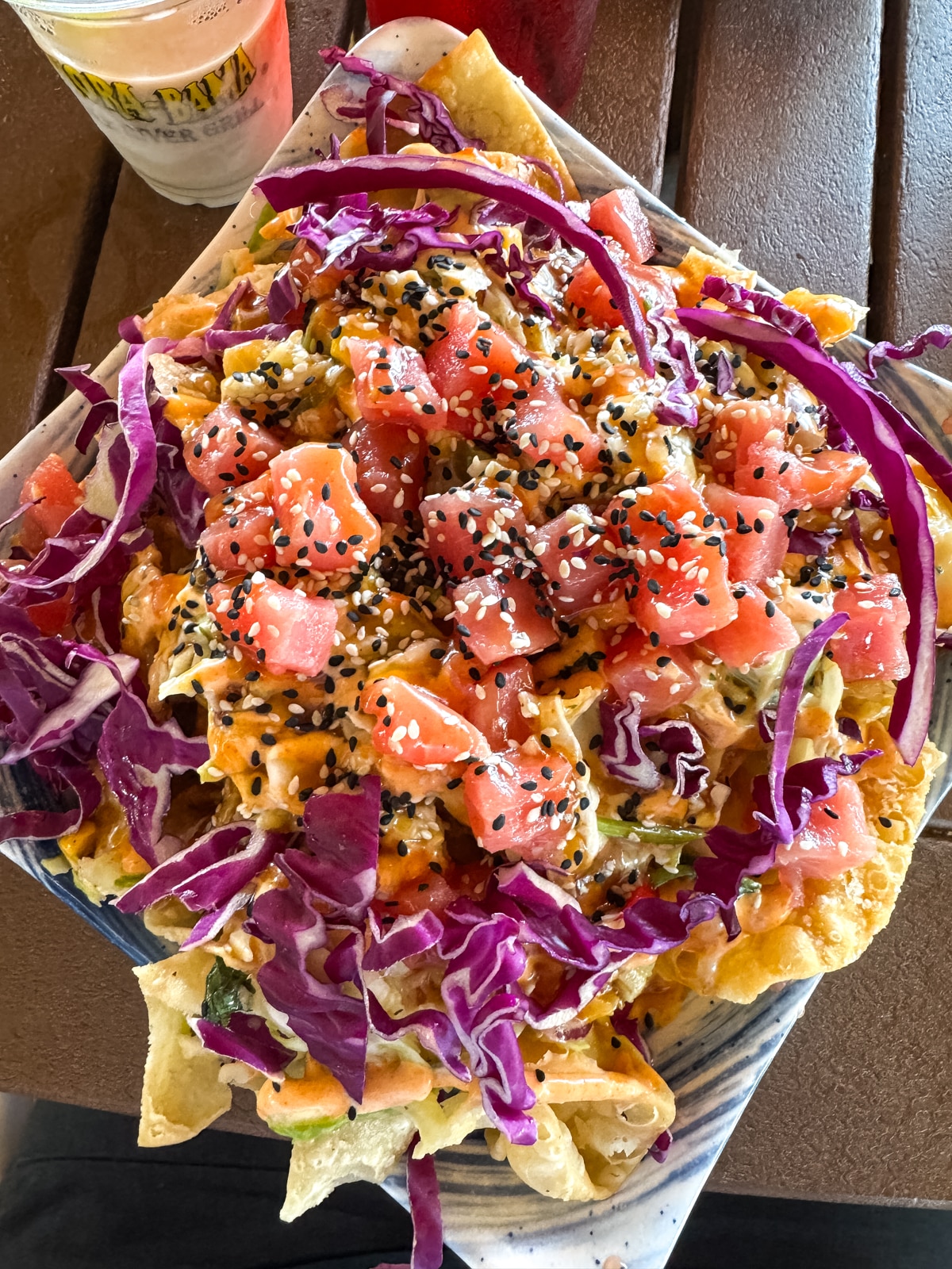 A plate of nachos topped with diced raw fish, shredded purple cabbage, sesame seeds, and a creamy orange sauce on a wooden table.