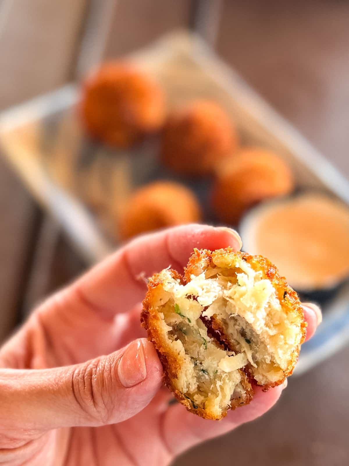 A hand holds a half-eaten fried ball revealing a cheesy, herbed filling; more fried balls and a dipping sauce are on a tray in the background.