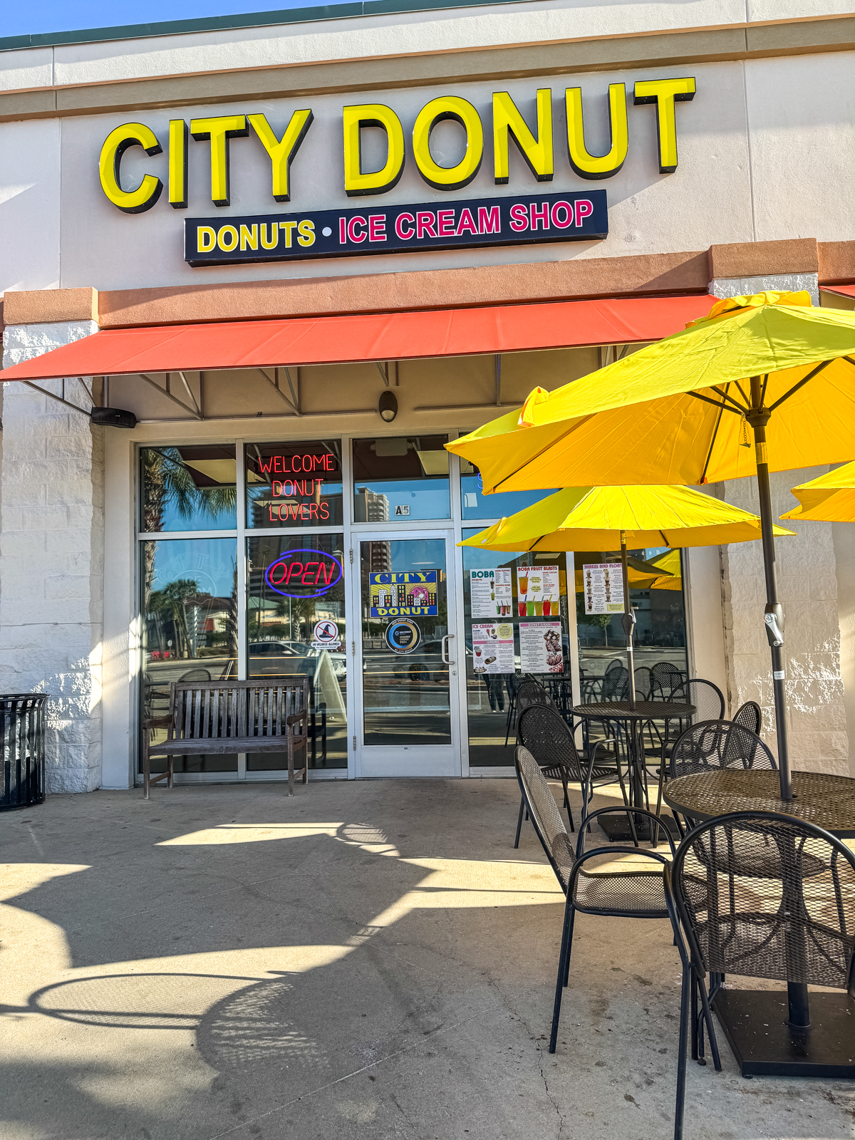 Outdoor seating area with yellow umbrellas in front of City Donut, a donuts and ice cream shop, with a glass entrance and various signs on the door and windows.