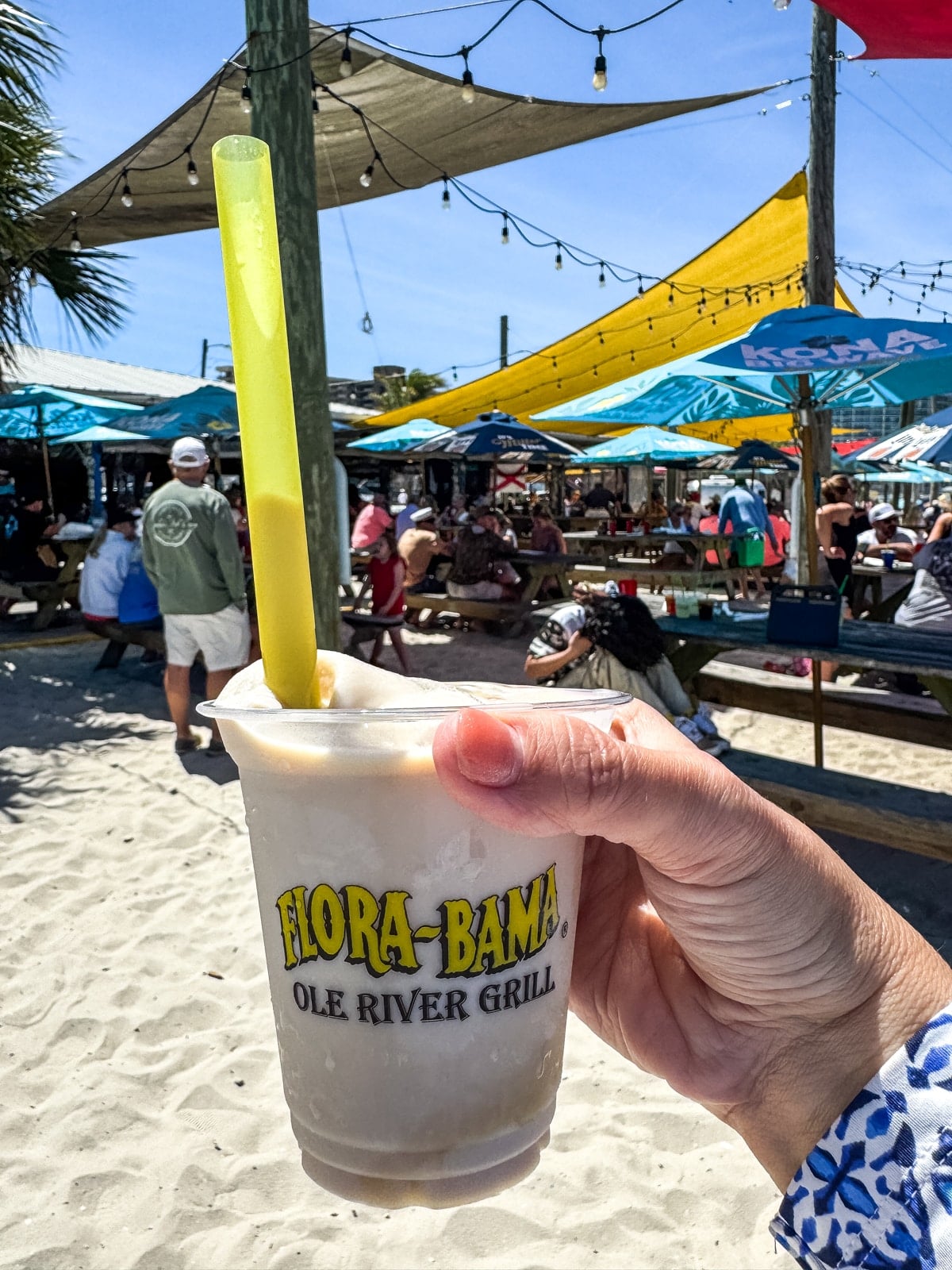 A hand holds a frozen drink in a Flora-Bama Ole River Grill cup with a yellow straw at an outdoor beachside restaurant with people, umbrellas, and string lights.