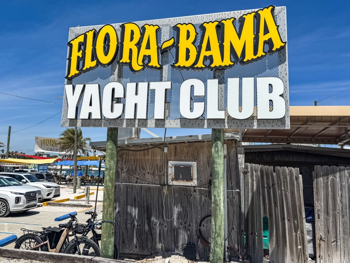 Large sign reading "Flora-Bama Yacht Club" stands above a wooden fence with bicycles and parked cars visible nearby under a clear blue sky.