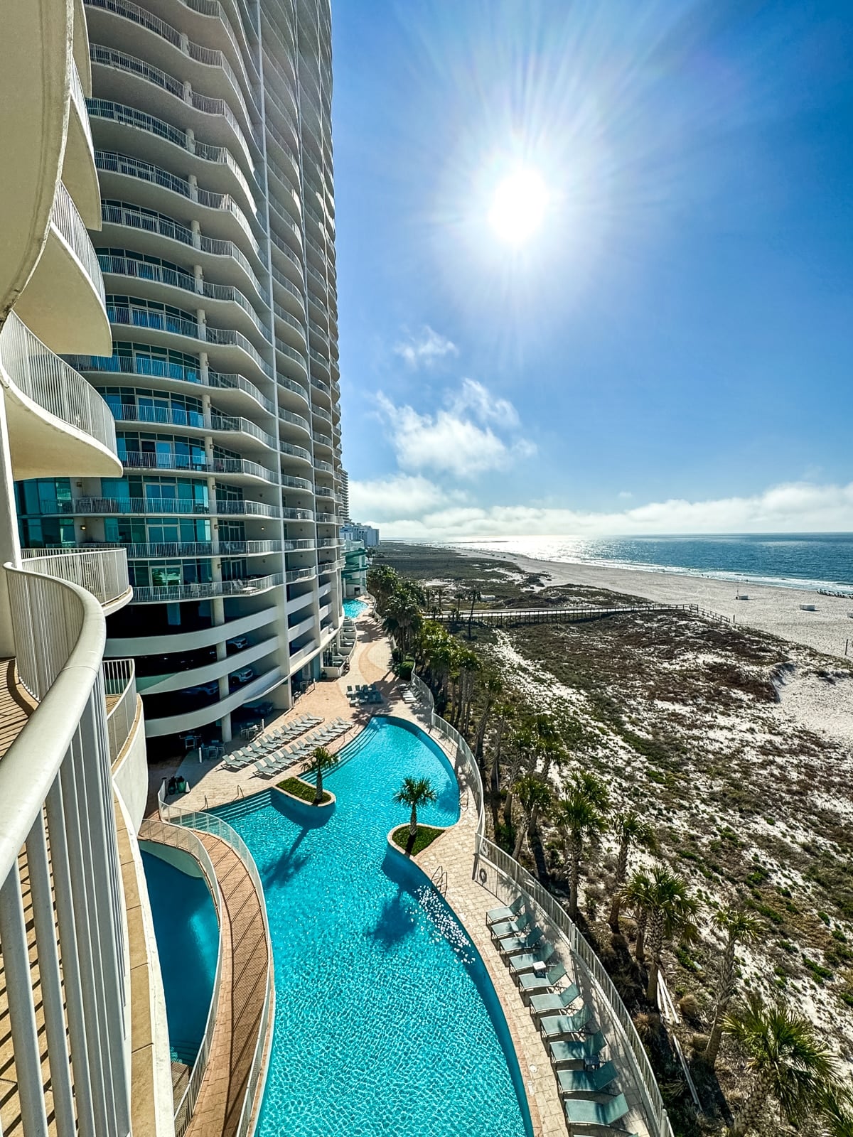 High-rise beachfront hotel with a long outdoor pool, sun loungers, palm trees, and a sandy beach under a bright, sunny sky.