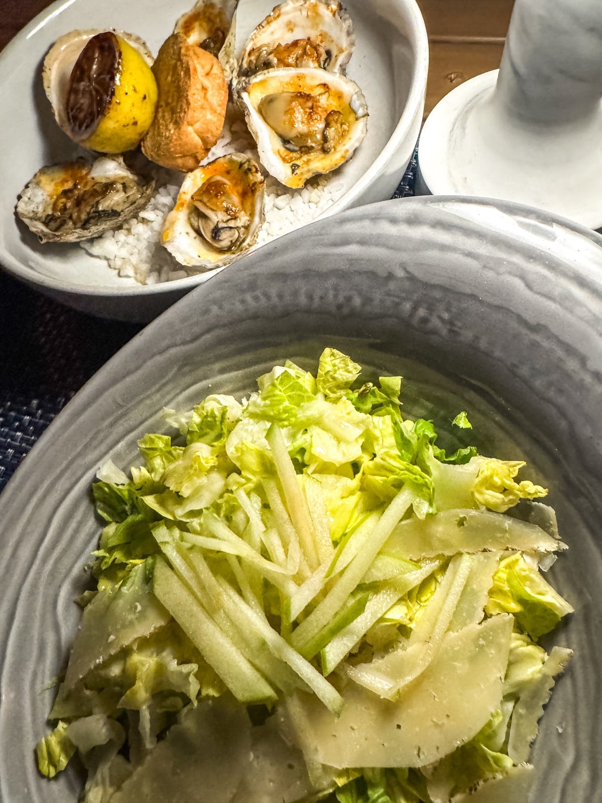 A bowl of green salad with shaved cheese and julienned apples in the foreground, with a plate of baked oysters and lemon in the background.
