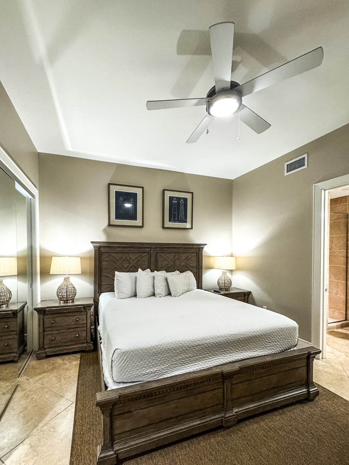 A bedroom with a large wooden bed, white bedding, two bedside tables with lamps, a ceiling fan, mirrored closet doors, and neutral-colored walls.