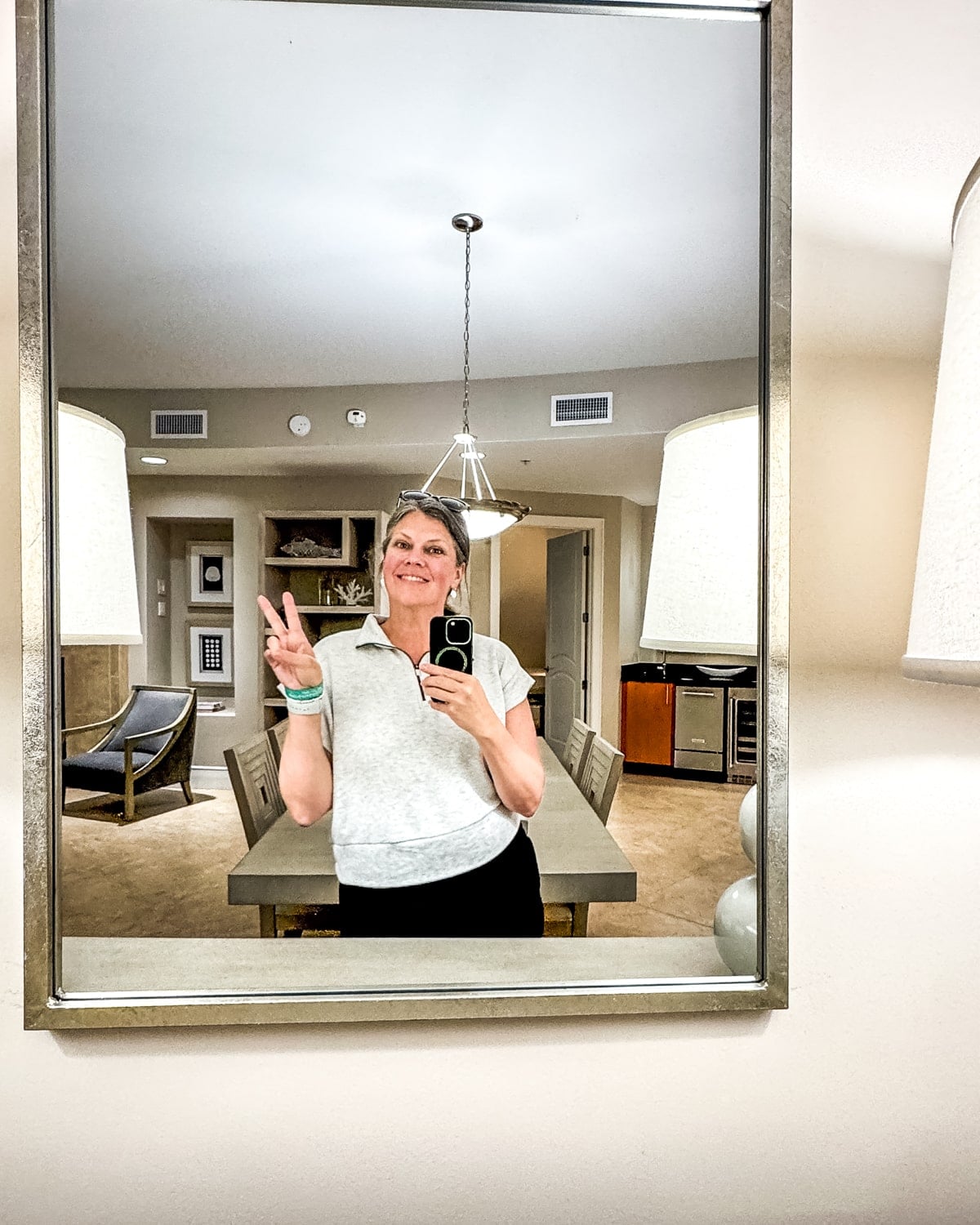 A person stands in front of a mirror, holding a phone and making a peace sign, with a dining room and furniture visible in the background.