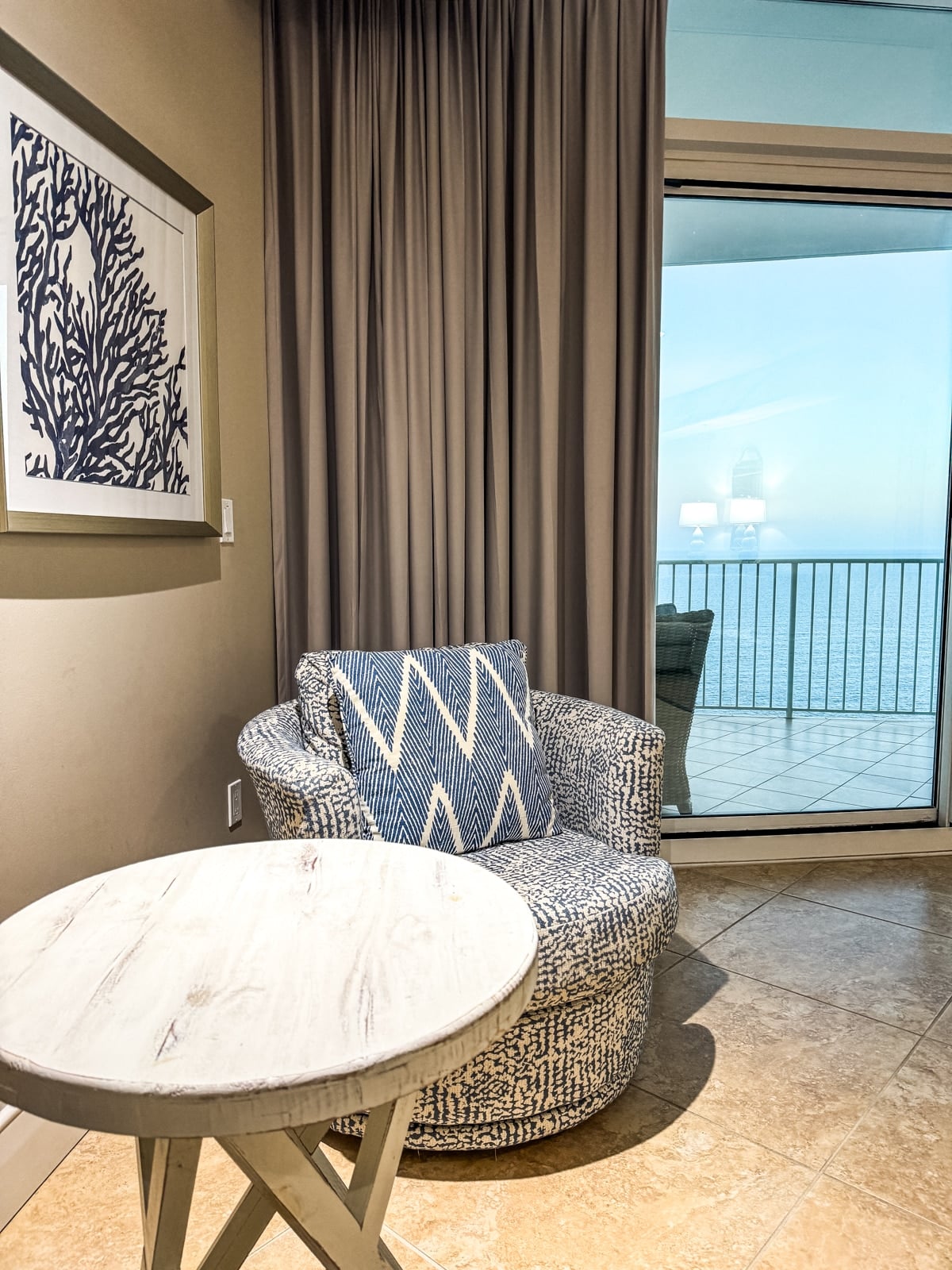 A patterned armchair and round wooden table sit by a curtained window overlooking a balcony with an ocean view. A framed coral artwork hangs on the wall.