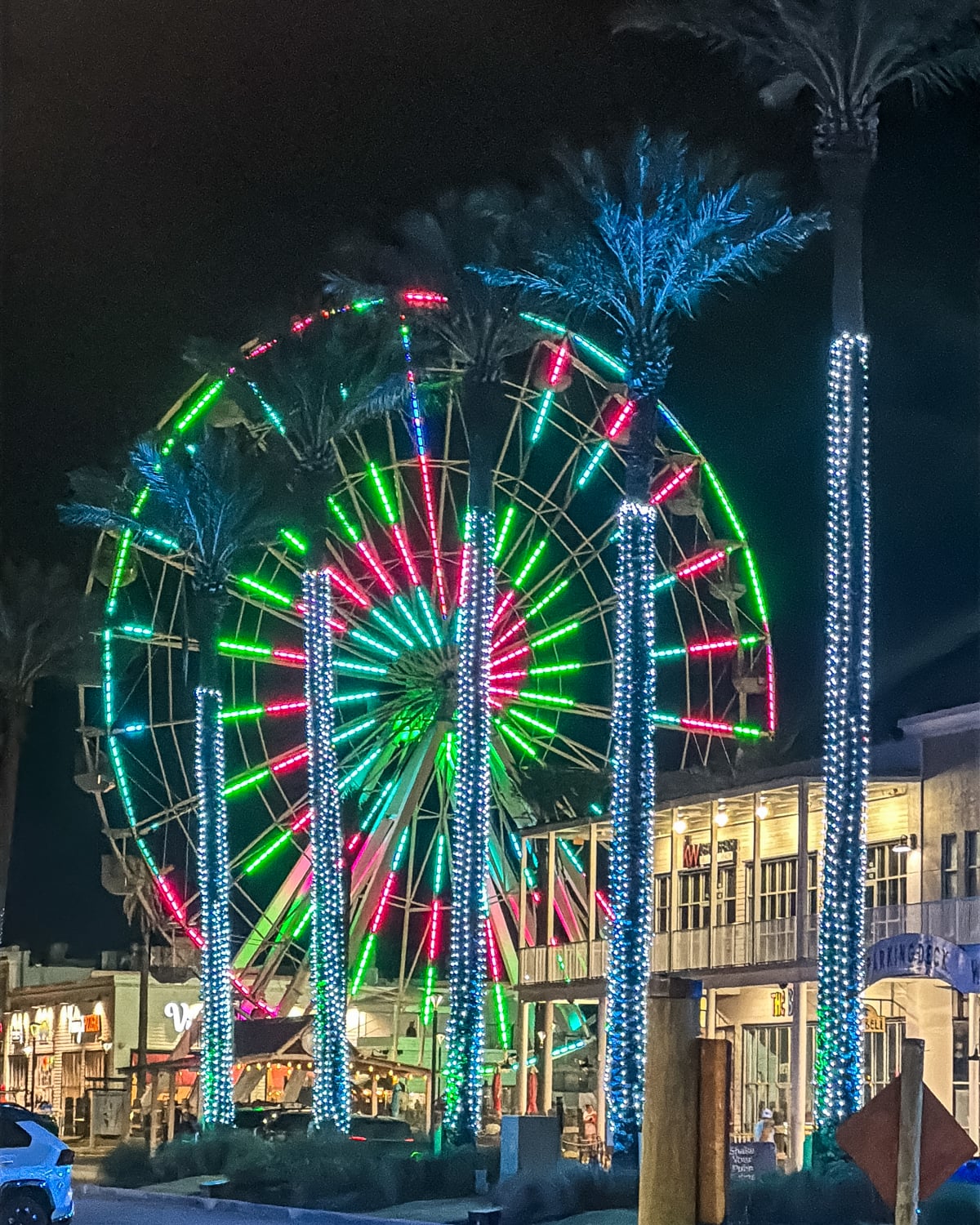 A Ferris wheel with red and green lights is illuminated at night behind palm trees wrapped in blue lights, near shops and restaurants.