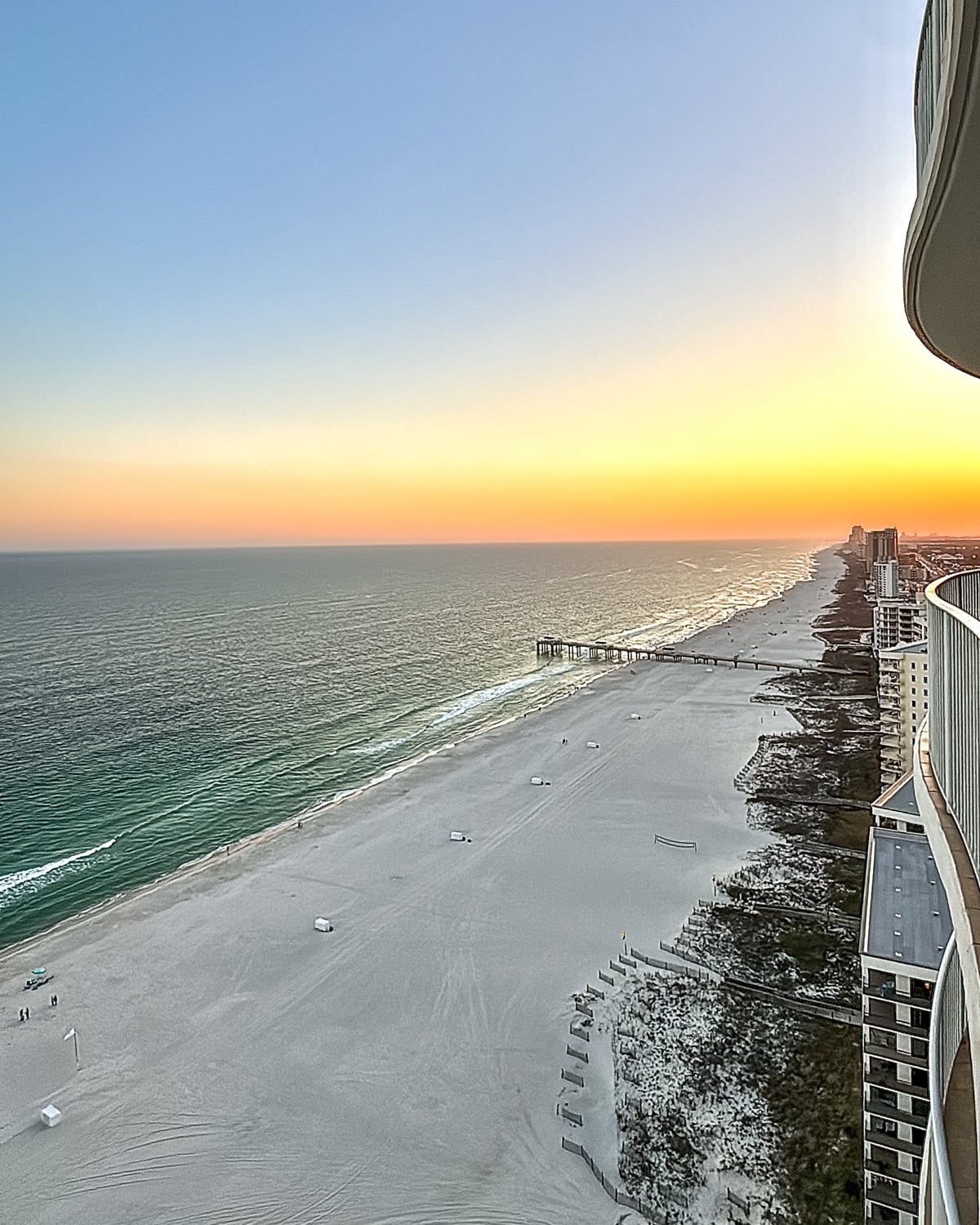 High-angle view of a sandy beach, calm ocean waves, a pier extending into the water, and a distant sunset, seen from a tall building balcony.