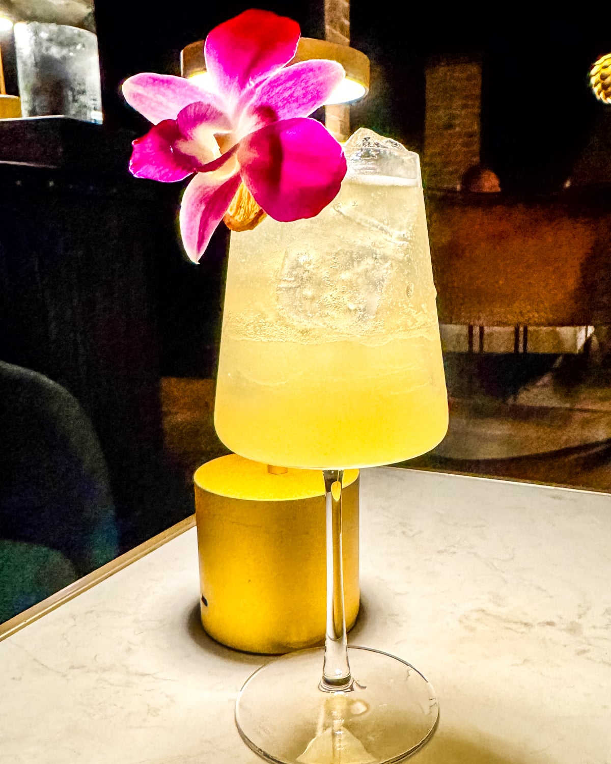 A yellow cocktail with ice is served in a stemmed glass, garnished with a pink and white orchid flower, placed on a marble table.