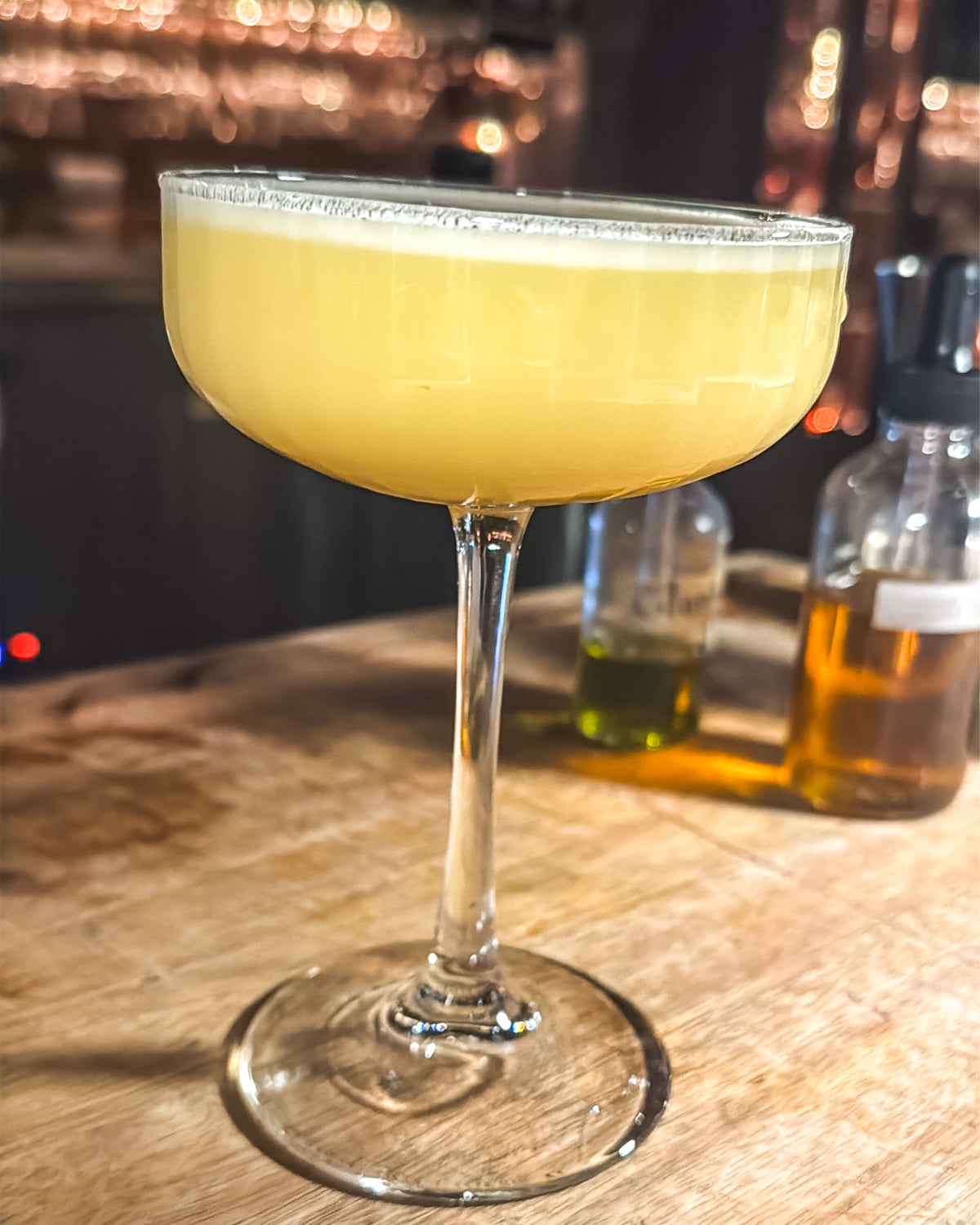 A coupe glass filled with a yellow cocktail sits on a wooden bar next to small bottles containing clear and green liquids.