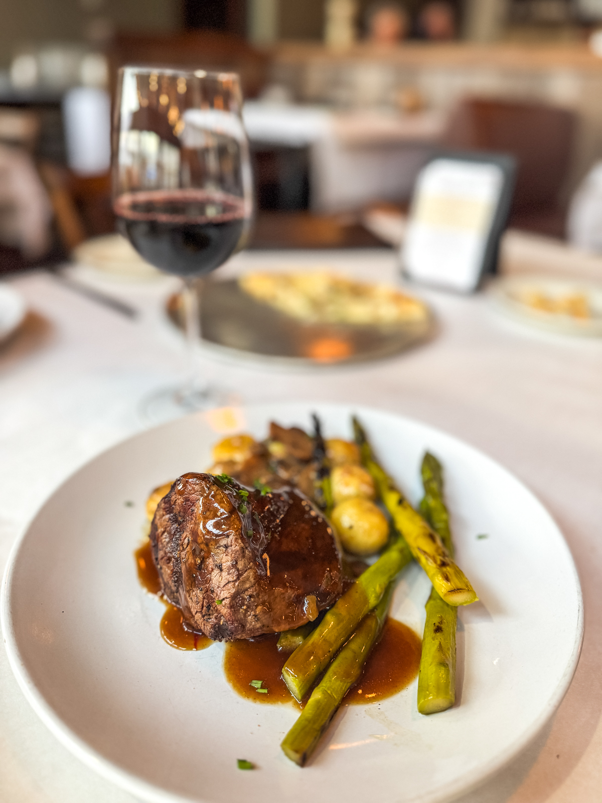 A plate of grilled steak with asparagus and potatoes, served with brown sauce, sits on a white tablecloth beside a glass of red wine in a restaurant setting.