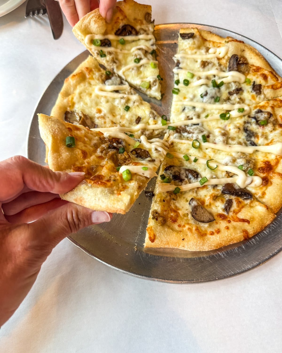 A hand reaches to take a slice of thin-crust pizza topped with mushrooms, green onions, cheese, and a drizzle of white sauce, served on a metal tray.